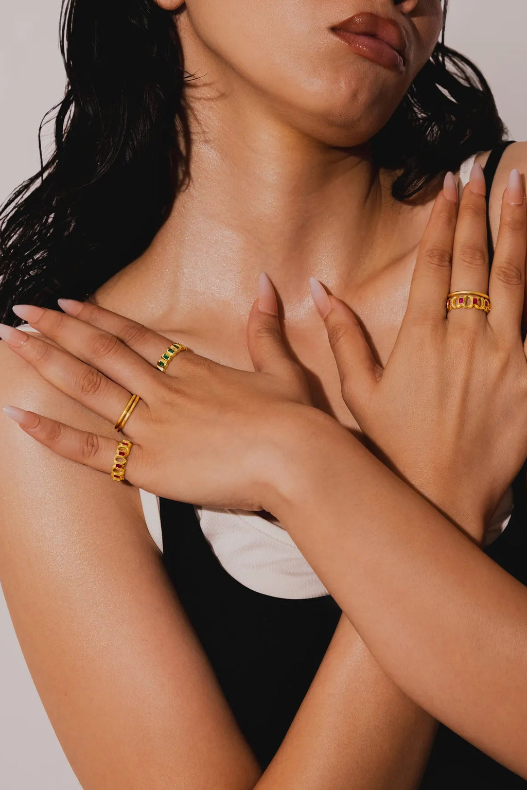 A close-up lifestyle shot of a woman wearing multiple gold rings on her hands and neck, posing against a neutral light gray background.