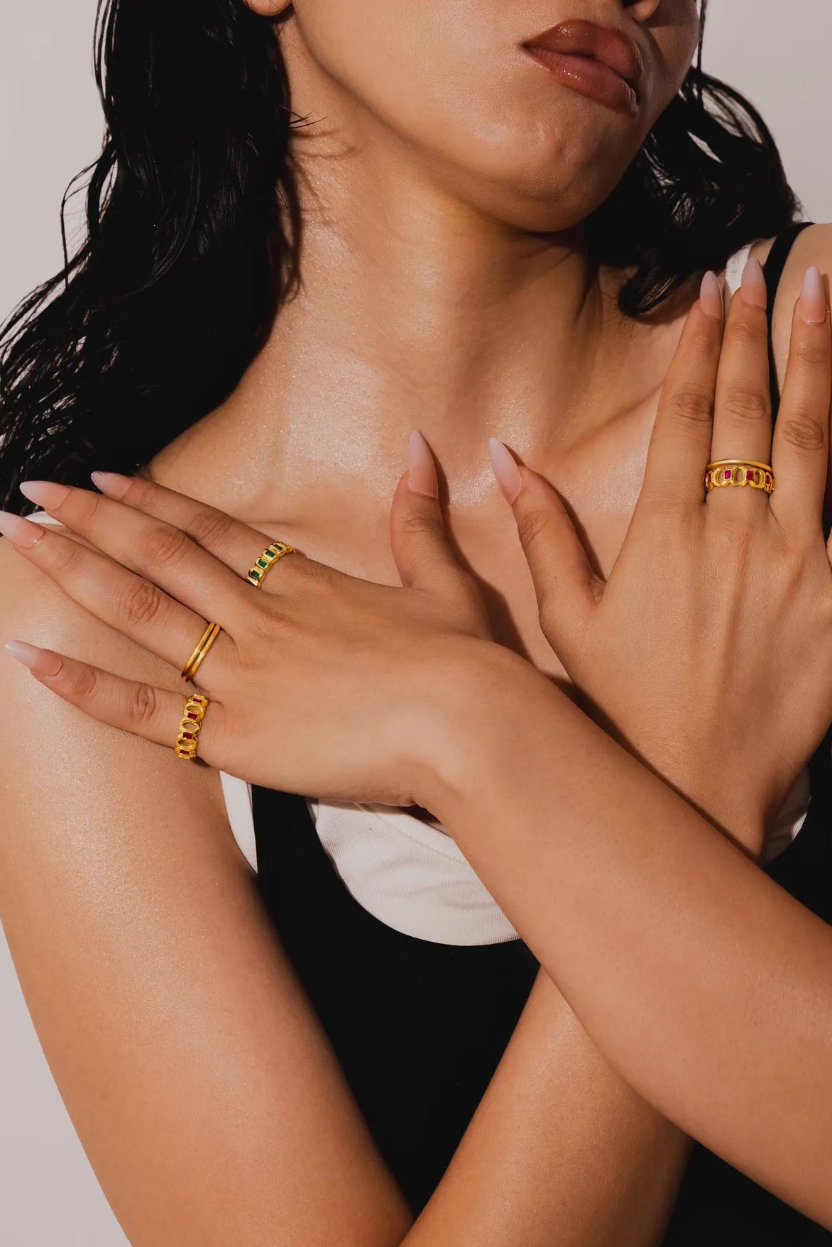 A close-up lifestyle shot of a woman wearing multiple gold rings on her hands and neck, posing against a neutral light gray background.