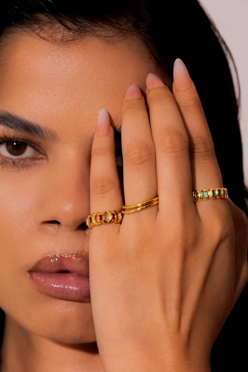 A close-up of a woman with dark hair wearing gold rings on her fingers, holding her hand near her face against a plain light background.
